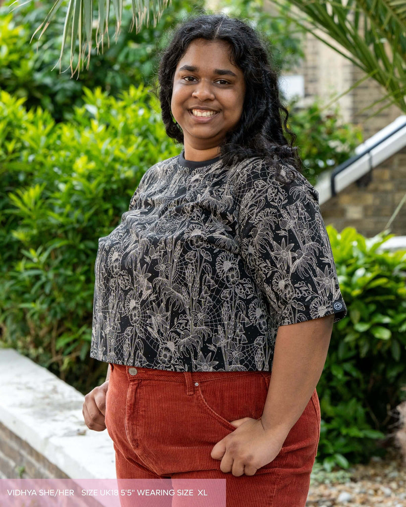 Vidhya, a lady with long curly black hair wearing the oversized boxy crop tee in a black and white spiderweb floral print. She is facing left looking towards the camera smiling with her hands in her orange corduroy trousers. 