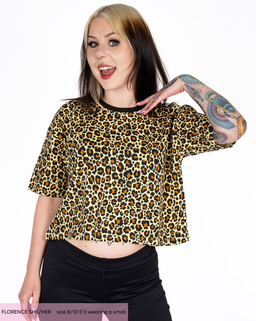 Florence, an alternative tattooed woman wearing a neutral leopard print crop top with contrasting black neck line. She's got matching coloured dyed hair in blonde and black. She's paired the top with black flared leggings. 
