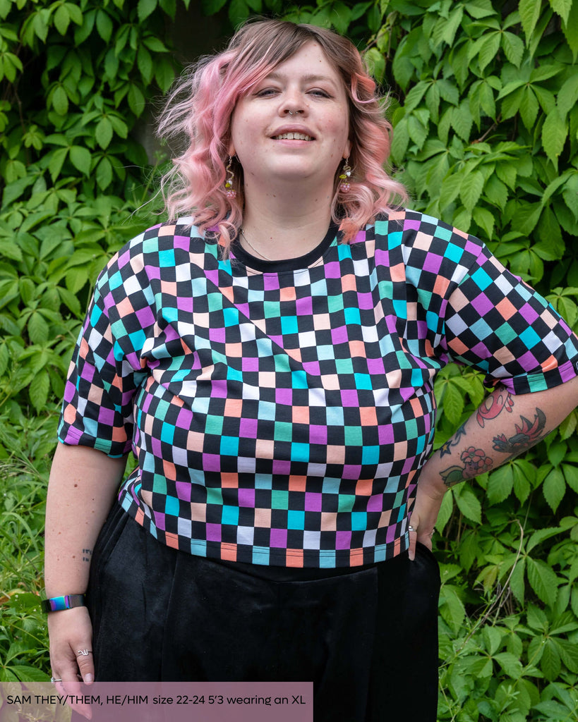 Sam strikes a confident pose with one hand on their hip, showcasing the cropped cut and bold pastel checkerboard print of the Rebel Romance tee. The look is playful and expressive, set against a natural leafy wall.