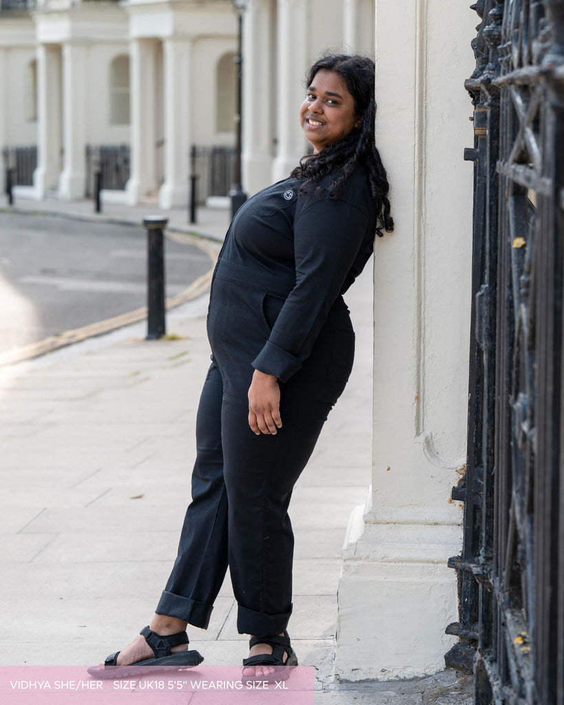 Woman in a black boiler suit standing leaning against a white building with a decorative black gate. She is facing her head to camera smiling. 
