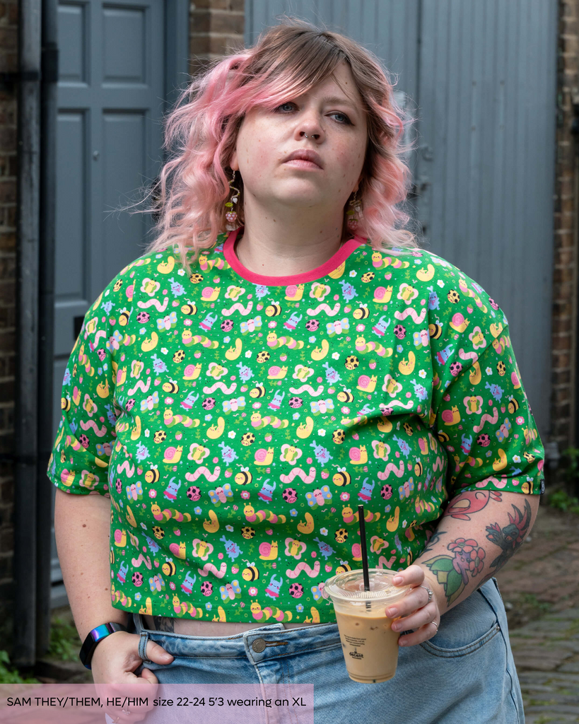 Sam, a tattooed non binary model with pink bobbed hair wearing the dipstick bugs crop top that has a contrasting pink neckline with light blue denim jeans. The print is a bright green base with adorable illustrated bees, ladybugs, slugs, butterfly, stag beetles, worms and caterpillars with florals and decorative doodles from dipstick. 