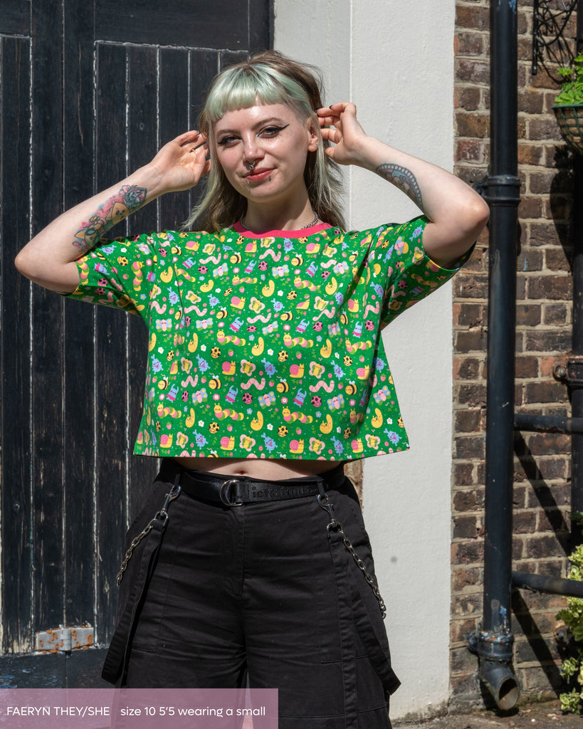 Faeryn, a non binary femme person wearing the dipstick bugs crop top with oversized black cargo trousers smiling stood outside. The top is a bright green base with pastel coloured kawaii bugs all over with a pink contrasting neck trim. 