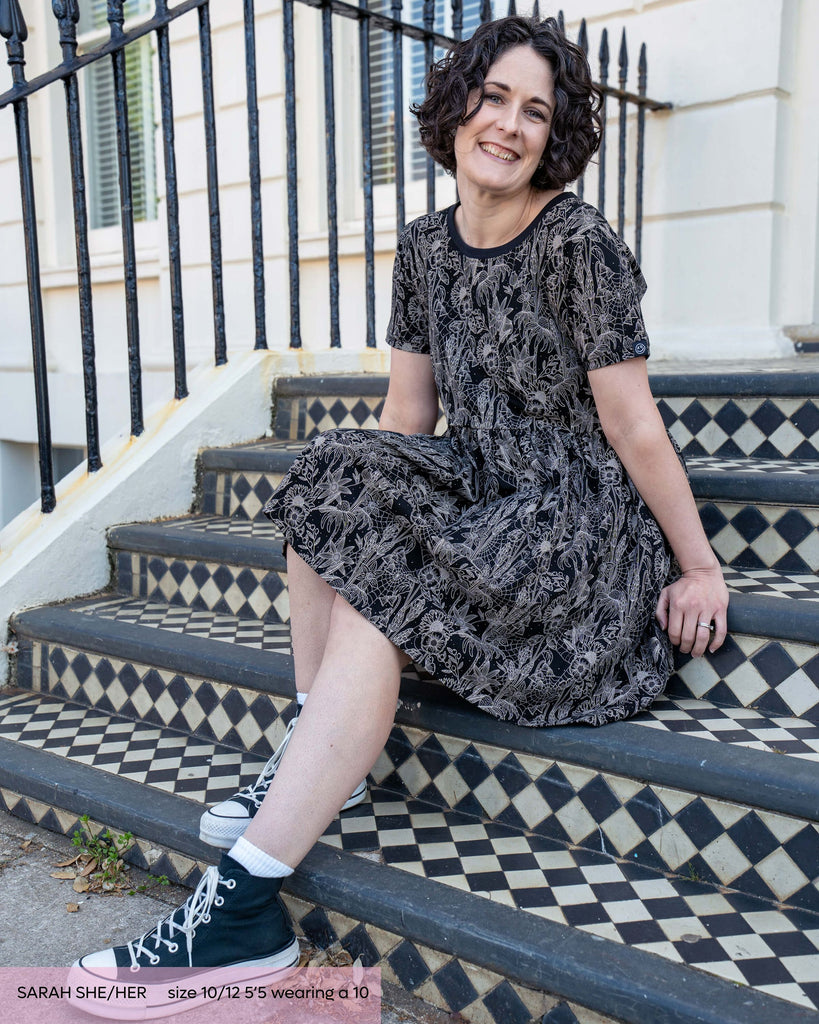 Sarah, a woman with short black curly hair wearing the black floral cobweb smock dress with black trainers. She is sat smiling on a set of black and white steps outside a white brick and black gated house. The smock dress print is a black base with white floral outlines, cobwebs and sparkles. 