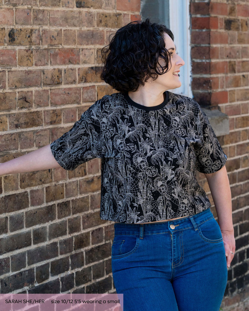 Sarah, a woman with black curly short hair smiling leaning on a wall looking right. She is wearing an oversized crop tshirt in black with a white cobweb floral print paired with blue denim jeans. 