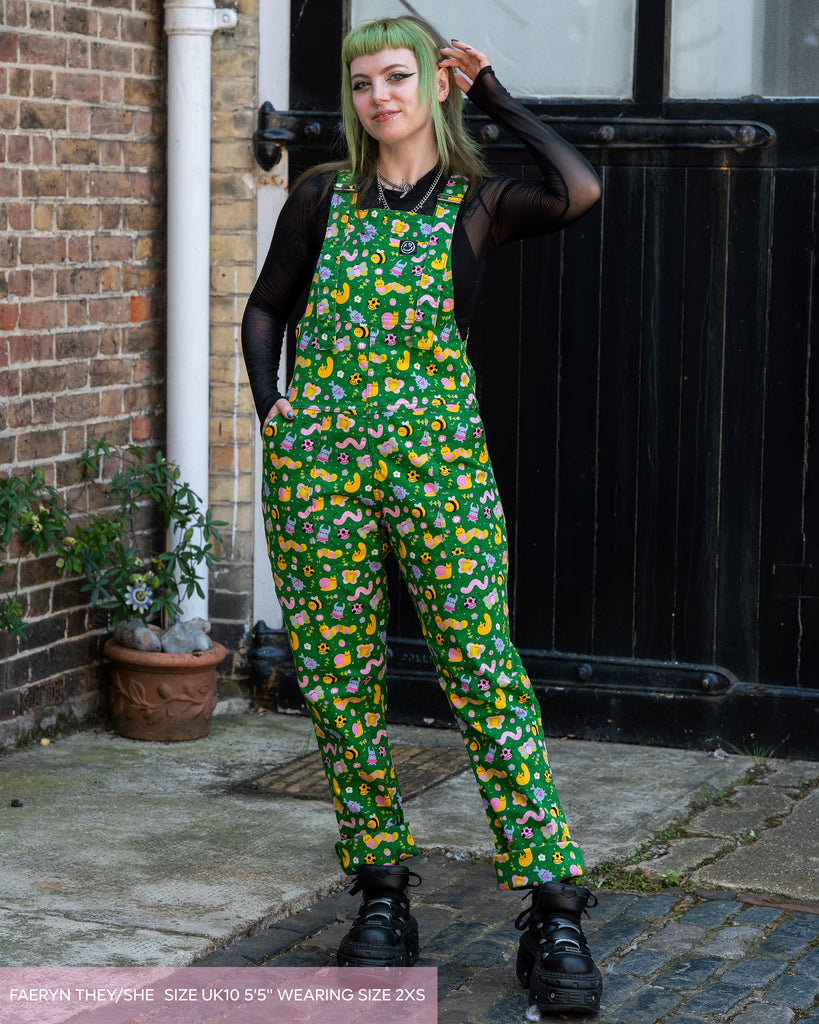 Faeryn wears Rebel Romance x Dipstick Store Bugs Stretch Dungarees in bright green with a cute bug and flower print. They stand outside against a black wooden door and brick wall, smiling with one hand in their pocket. Alternative slow fashion from a UK brand celebrating unique style.