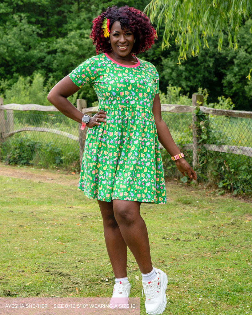 Ayesha, a brown and red curly haired woman wearing the dipstick bugs smock dress with matching hair bows, pink beaded bracelet and white hello kitty trainers. She is stood smiling in a grassy area by a pond with a weeping willow tree leaning over. The dress print is a bright green colour with adorable bugs all over illustrated by Dipstick, it has a bright pink contrasting neck trim. 