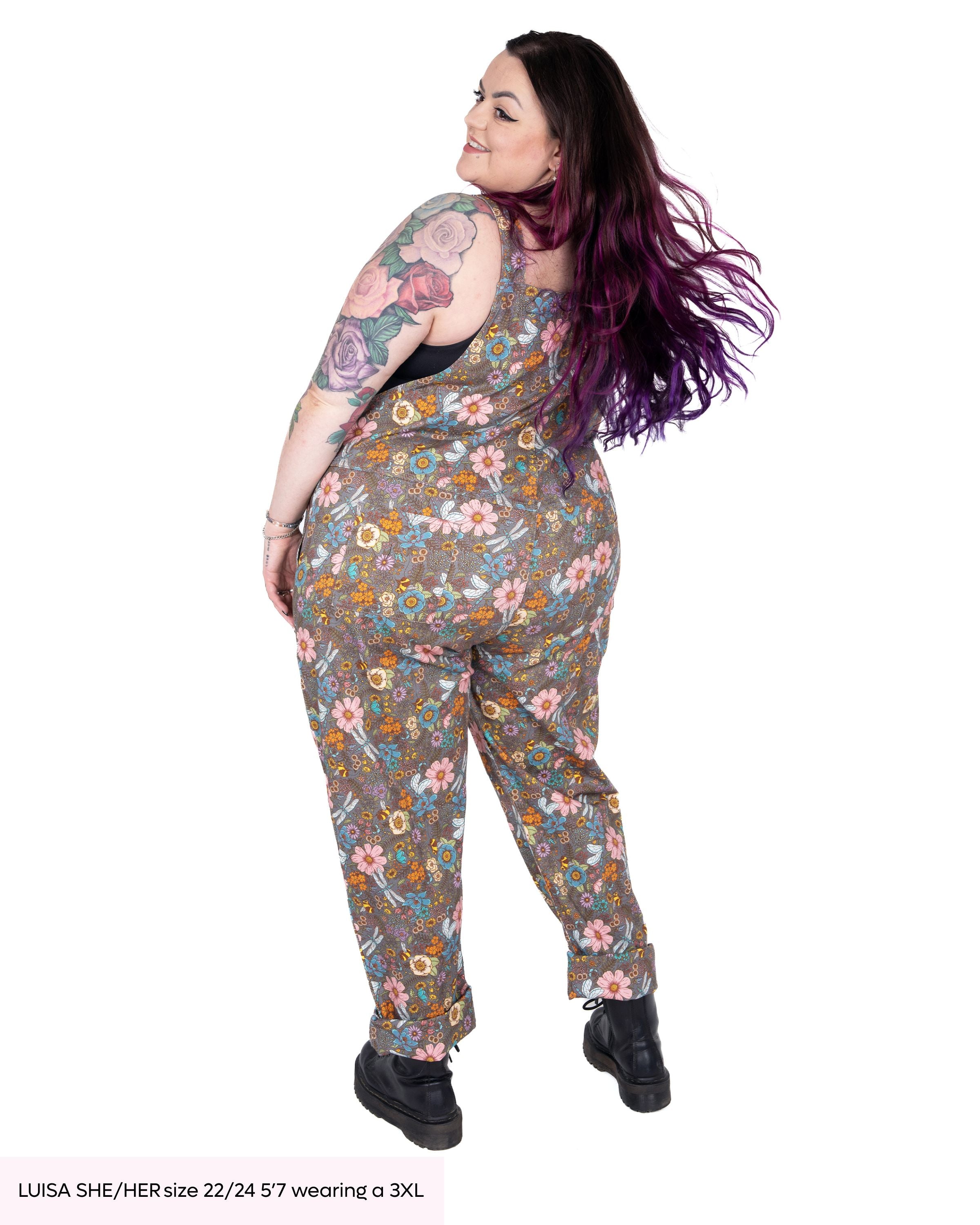 Luisa, a tattooed model with purple and pink hair wearing the rebel romance cottagecore dungarees with black platform boots. She is facing away flicking her long colourful hair over her shoulder in a white studio background. The dungarees print is a warm grey base with various coloured florals, bees, dragonflies and butterflies.