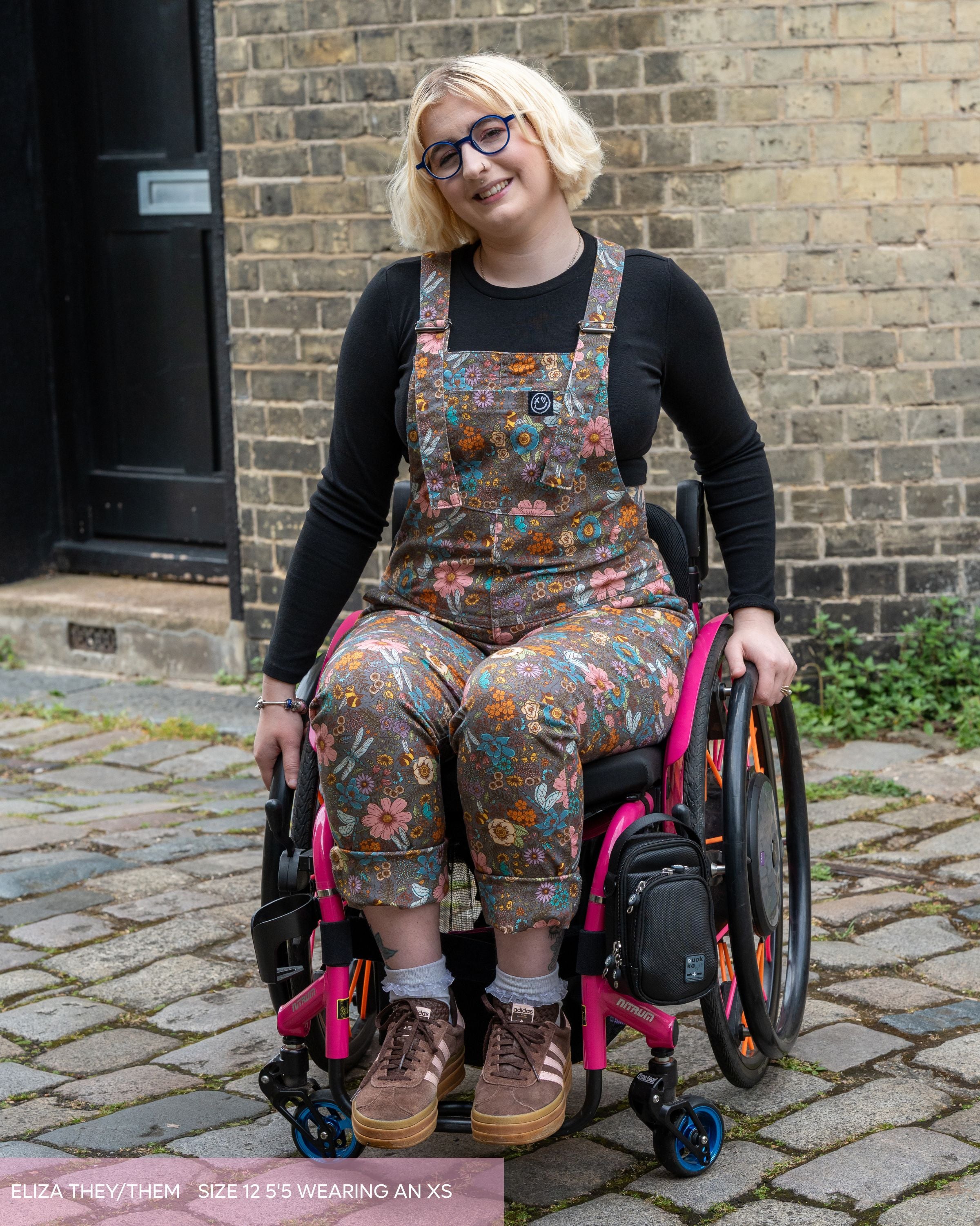 Eliza, a model with blonde cropped hair wearing rebel romance cottagecore dungarees, brown trainers and black long sleeve tshirt. They are leaning forward smiling in their wheelchair with pink accents. The dungaree print is a warm grey brown base with various pastel colourful flowers, bees, dragonflies and butterflies. 
