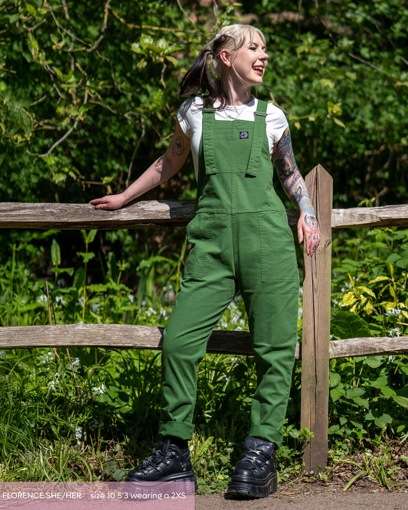 Florence, a tattooed model with ghost roots hair laughing looking right leaning on a wooden fence. She is wearing the forest green rebel romance dungarees with a white short sleeve crop top and black platform trainers. She is stood outside with a wooden fence and park area behind her. 