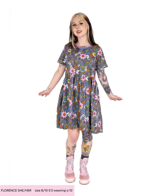 Florence, a tattooed alternative model with dyed bleach blonde and black hair wearing the cottagecore smock dress and pink platform boots. The dress is a denim blue base with contrasting purple neck trim, the print features various colourful florals, bees, dragonflies and butterflies. 