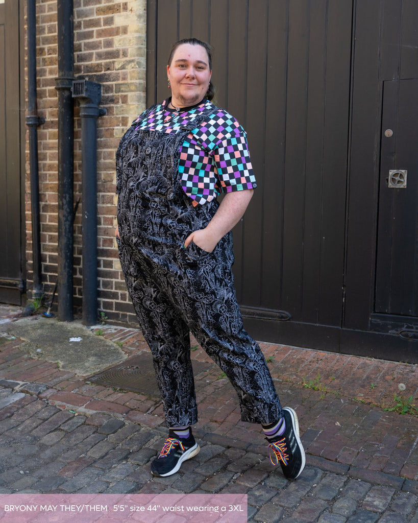 Bryony May, a non binary person wearing the rebel romance black floral cobweb dungarees with the rebel romance checkerboard crop top and black rainbow laced trainers. They are taking a step left smiling to camera with both hands in the pockets. The print is a black base with white floral outlines with cobwebs. 