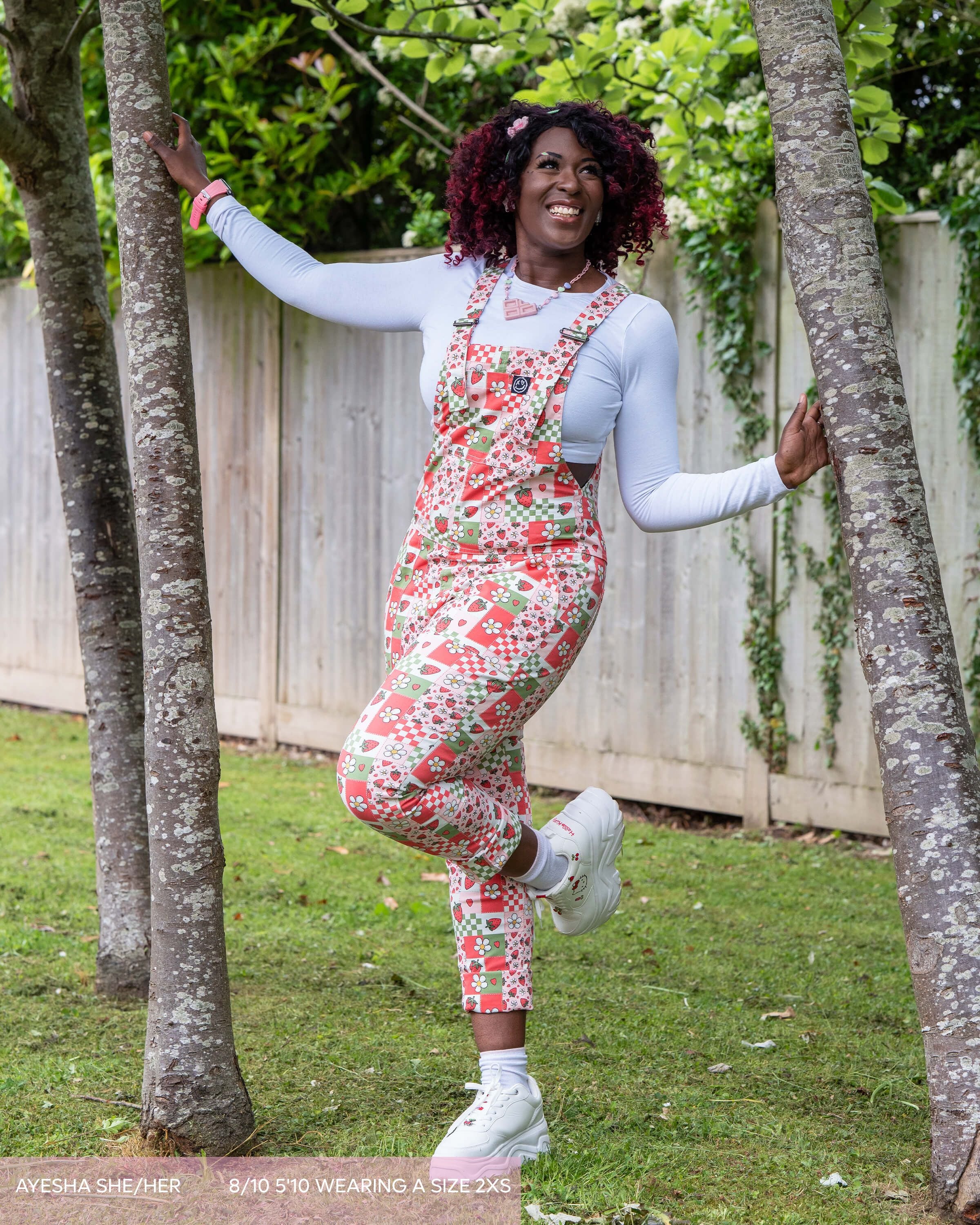 Ayesha, a kawaii style woman with red and black curly hair wearing the strawberry picnic dungarees with a candy necklace, white long sleeve tshirt and white hello kitty trainers. She is smiling off to the right leaning between two trees. The print features strawberries, blossom flowers and checkerboard in green and pink. 