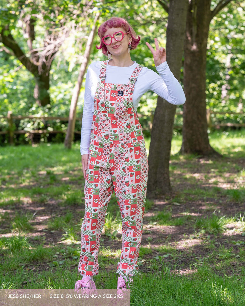 Jess, a pink haired woman with sparkly makeup wearing the strawberry picnic dungarees with a white long sleeve, pink heart glasses and pink heeled crocs. She is posing facing forward smiling holding up a peace sign. The dungaree print is a pastel pink, white and sage green with strawberries and blossoms in a checkerboard style.
