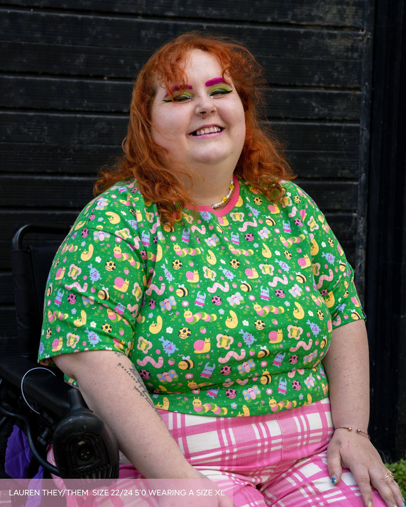 Lauren, a non binary model with ginger curly hair and brightly coloured makeup wearing the dipstick bugs crop top tshirt with pink checkerboard trousers. The top has a pink contrasting neck trim with a bright green base and illustrated cute bugs all over.