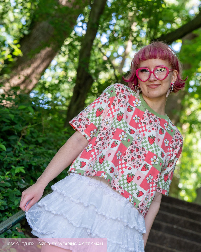 Jess, a sparkly pink human wearing the strawberry picnic crop top with white ruffle shorts smiling stood outside on some steps. She has pink fairy themed makeup, pink bobbed hair and pink heart glasses. The top print is a checkerboard sage green, strawberries, pink blossoms and more.