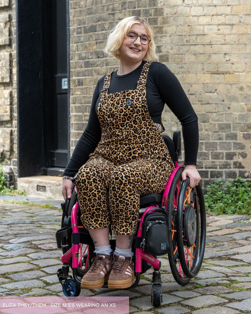 Eliza, a non binary disabled model with blonde cropped hair and tortoiseshell glasses. They are wearing a black longsleeve tshirt, brown trainers and the rebel romance neutral leopard print dungarees. 