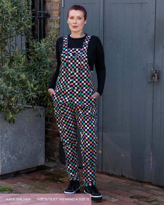 Katie, a tattooed woman wearing a colourful checkerboard patterned dungarees in colour block colours of blue, orange, purple, green, blue and pastels. They've paired it with black trainers and a black long sleeve tshirt underneath standing in front of a grey door.