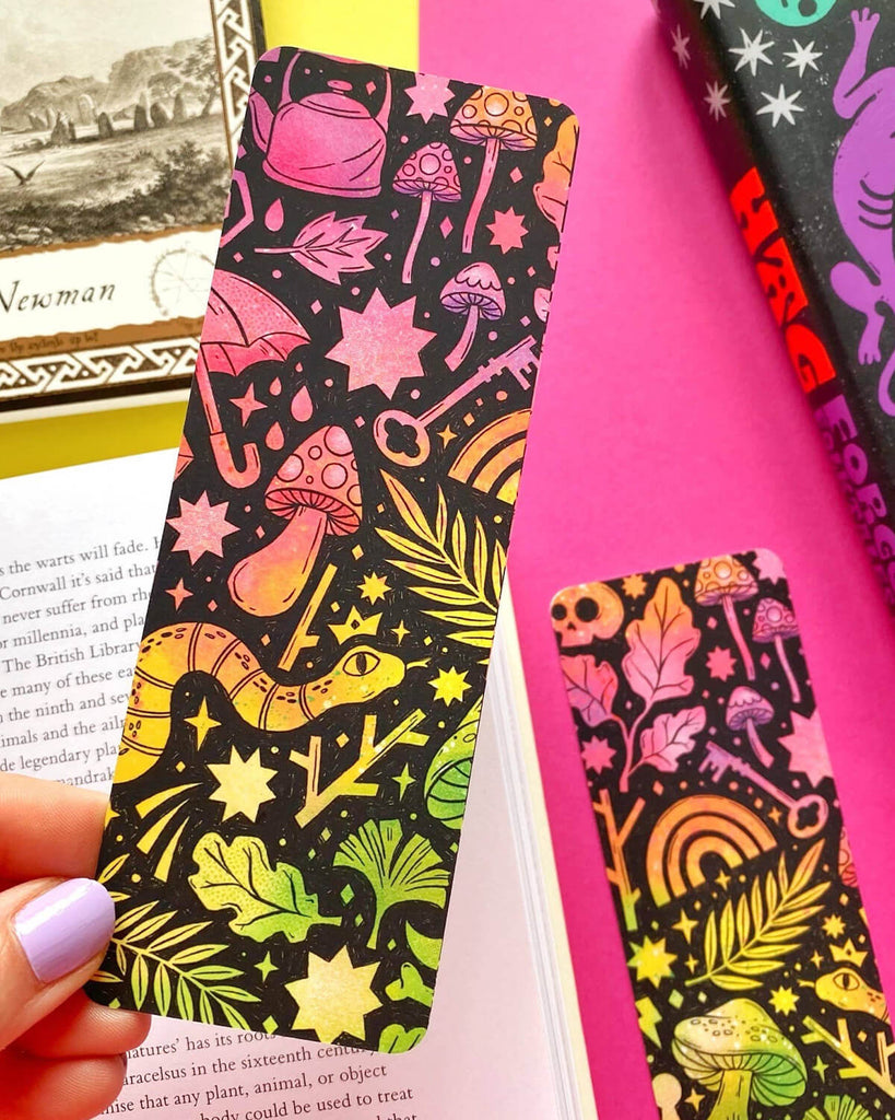 A hand with lilac nail polish holding a rectangular bookmark with a vibrant rainbow pattern of mushrooms, stars, leaves, snakes, and keys on a black background. A book and colourful stationery are in the background.