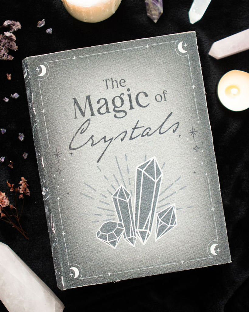 A mystical book-shaped storage box featuring a grey cover with the title “The Magic of Crystals” and illustrations of crystals, stars, moons, and celestial motifs. Shown open to reveal a secret compartment, perfect for storing jewellery, trinkets, or crystals. Ideal for gothic or witchy home decor.