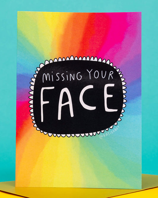 Bright and playful A6 greeting card by Katie Abey featuring a rainbow tie-dye background with a bold black doodle in the centre that reads “Missing Your Face” in quirky white hand lettering. The card is displayed standing on a colourful surface. Sold by Rebel Romance, a small UK brand celebrating alternative style and stationery.