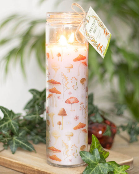  A lit mushroom-print tube candle with a glowing flame, tied with rustic twine and a decorative ‘Enchanted Forest’ tag, surrounded by ivy and greenery.