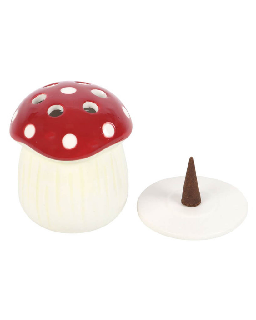 The mushroom incense cone holder on a white surface with a lit incense cone inside, releasing soft, fragrant smoke, with green leaves blurred in the background.