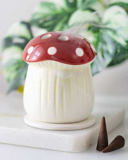 The mushroom incense cone holder on a white surface with a lit incense cone inside, releasing soft, fragrant smoke, with green leaves blurred in the background.
