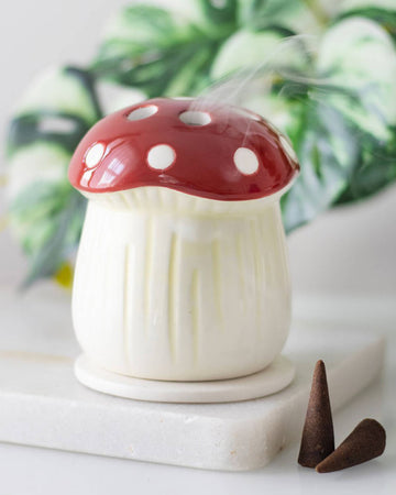 The mushroom incense cone holder on a white surface with a lit incense cone inside, releasing soft, fragrant smoke, with green leaves blurred in the background.