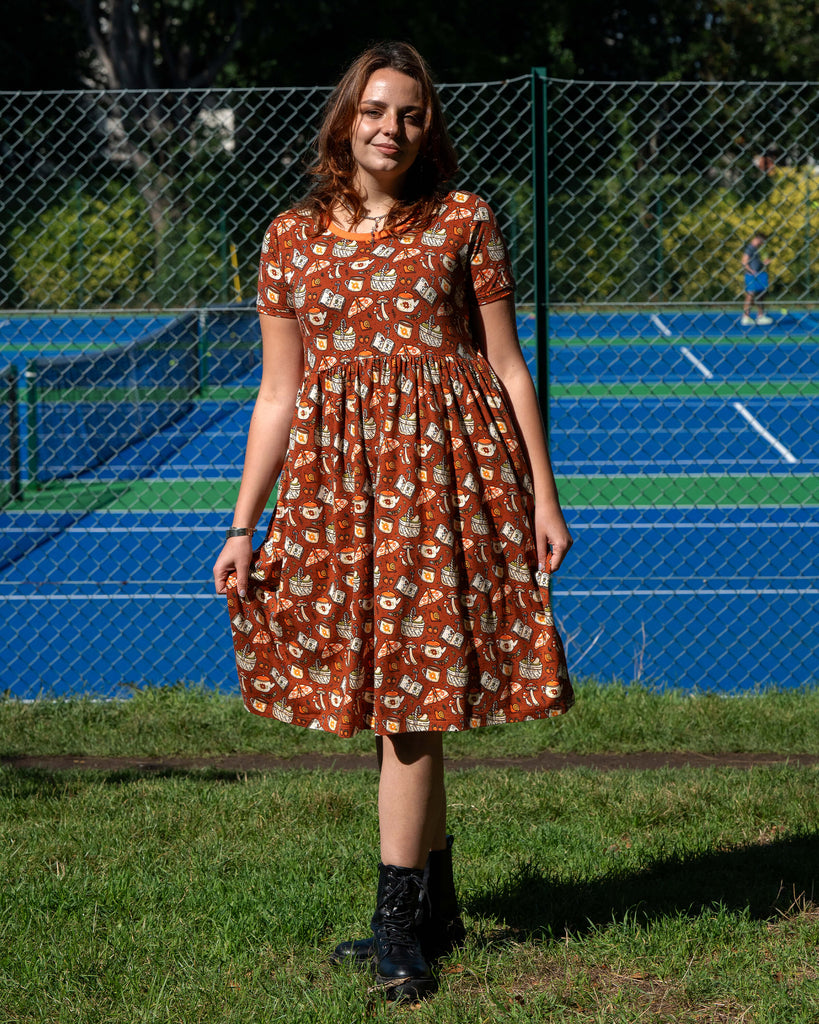 Model wearing the Mushroom Tea Party Midi Dress outdoors, styled with black boots. The rust-orange smock dress has a woodland-inspired pattern with mushrooms, moths, teapots, and books.