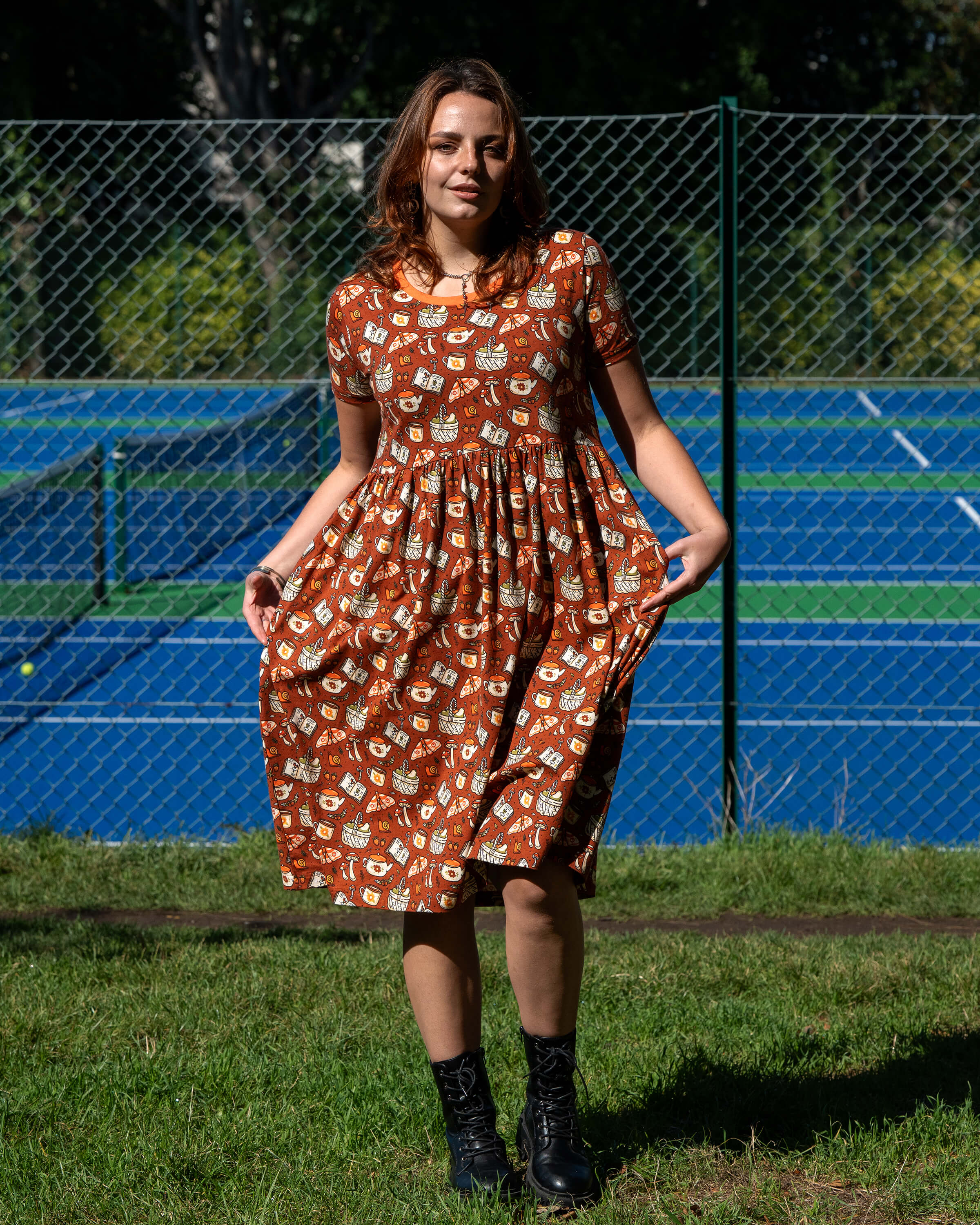 Model wearing the Mushroom Tea Party Midi Dress outdoors, styled with black boots. The rust-orange smock dress has a woodland-inspired pattern with mushrooms, moths, teapots, and books.