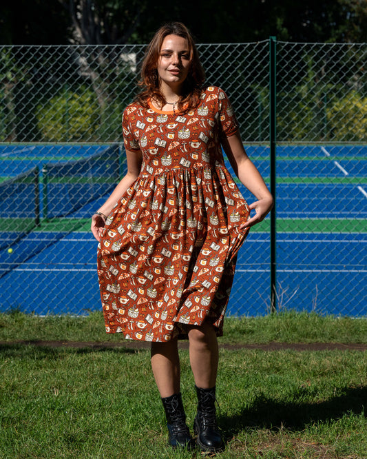Model wearing the Mushroom Tea Party Midi Dress outdoors, styled with black boots. The rust-orange smock dress has a woodland-inspired pattern with mushrooms, moths, teapots, and books.