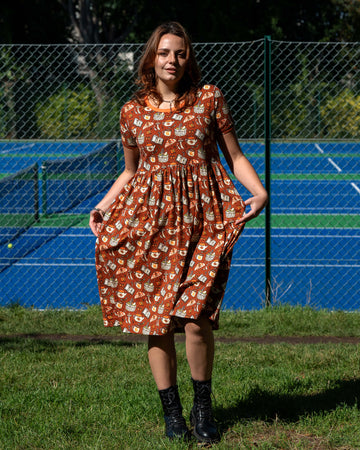 Model wearing the Mushroom Tea Party Midi Dress outdoors, styled with black boots. The rust-orange smock dress has a woodland-inspired pattern with mushrooms, moths, teapots, and books.