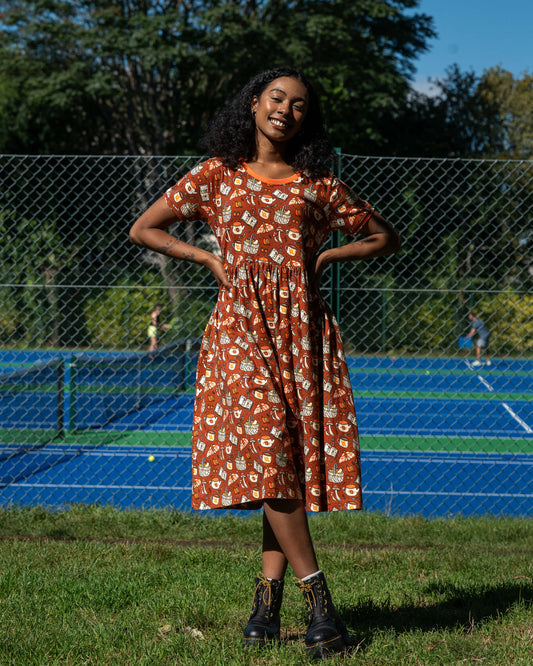 Model wearing the Mushroom Tea Party Midi Dress, smiling while adjusting her hair. The rust-orange smock dress features a whimsical woodland print of mushrooms, moths, books, and teapots.