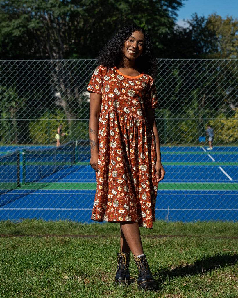 Model wearing the Mushroom Tea Party Midi Dress in rust-orange, smiling with hands on hips. The autumn-inspired print features mushrooms, moths, teapots, and books, styled with black lace-up boots.