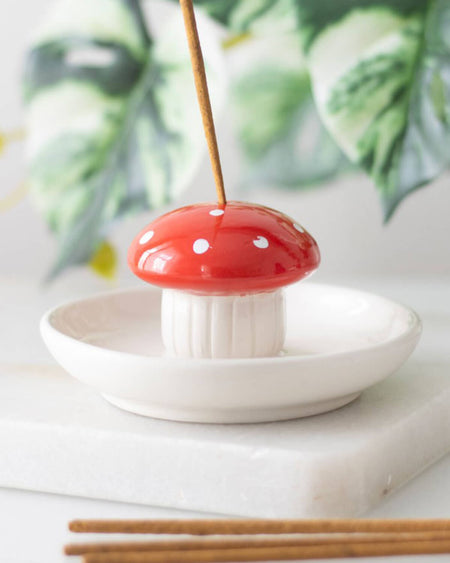 A ceramic mushroom-shaped incense holder with a red cap and white spots on a cream ceramic dish, holding an incense stick.