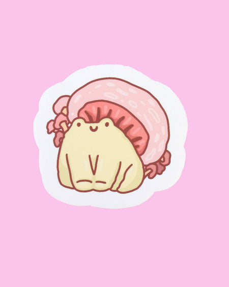 A glossy vinyl sticker featuring a hand-drawn illustration by Nimbob of a cute, chubby yellow frog sitting under a large pink mushroom cap. The mushroom has soft, rounded edges with a red underside, and tiny frills along its base. The frog has a tiny smiling face and stubby little legs. The sticker has a white border and is displayed on a pastel pink background with gold and silver star confetti. A perfect sticker for frog lovers, nature enthusiasts, and cottagecore fans!