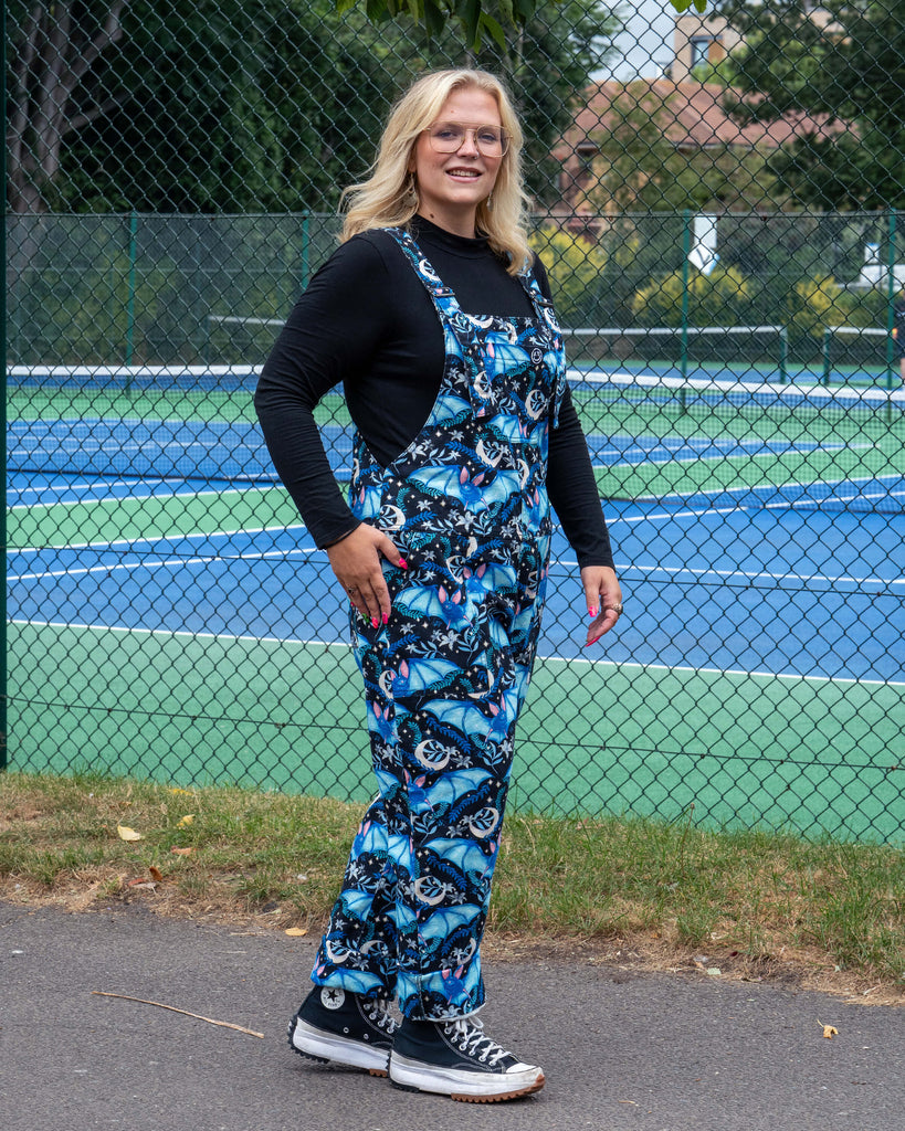Side view of Betty wearing Rebel Romance Night Flight Stretch Dungarees. The celestial bat print in blue tones stands out against the black fabric, styled over a black top with chunky Converse for an alternative everyday look.