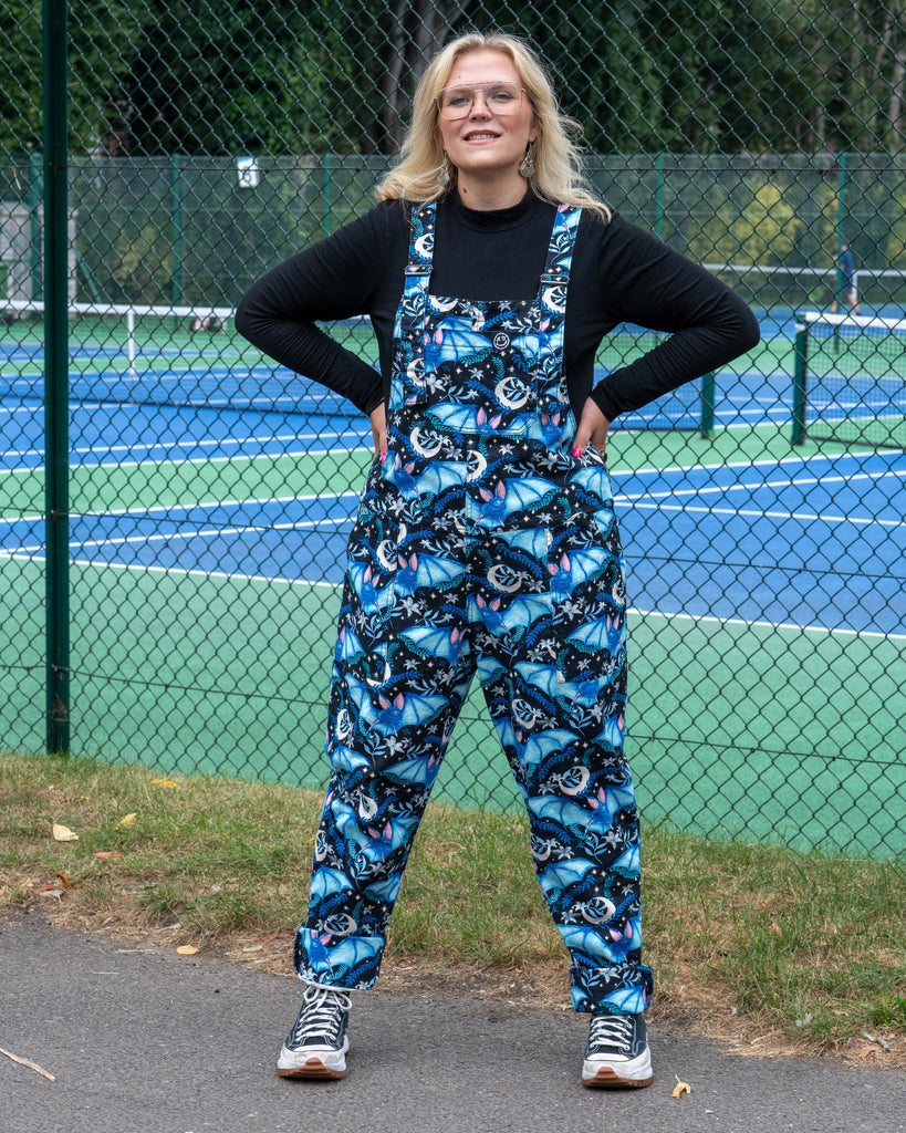 Betty poses in Rebel Romance Night Flight Stretch Dungarees, styled with rolled hems and chunky trainers. The celestial bat design in shades of blue makes these ethical dungarees a standout piece for alternative fashion lovers.