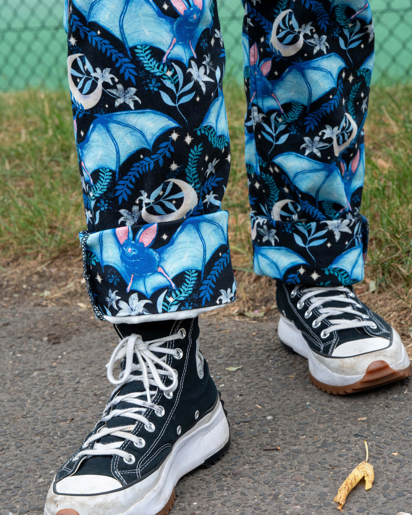 Close-up of the Rebel Romance Night Flight Dungarees showing the detailed blue bat and crescent moon print with floral accents. The rolled hems highlight the quality cotton twill fabric, styled with chunky black Converse trainers.