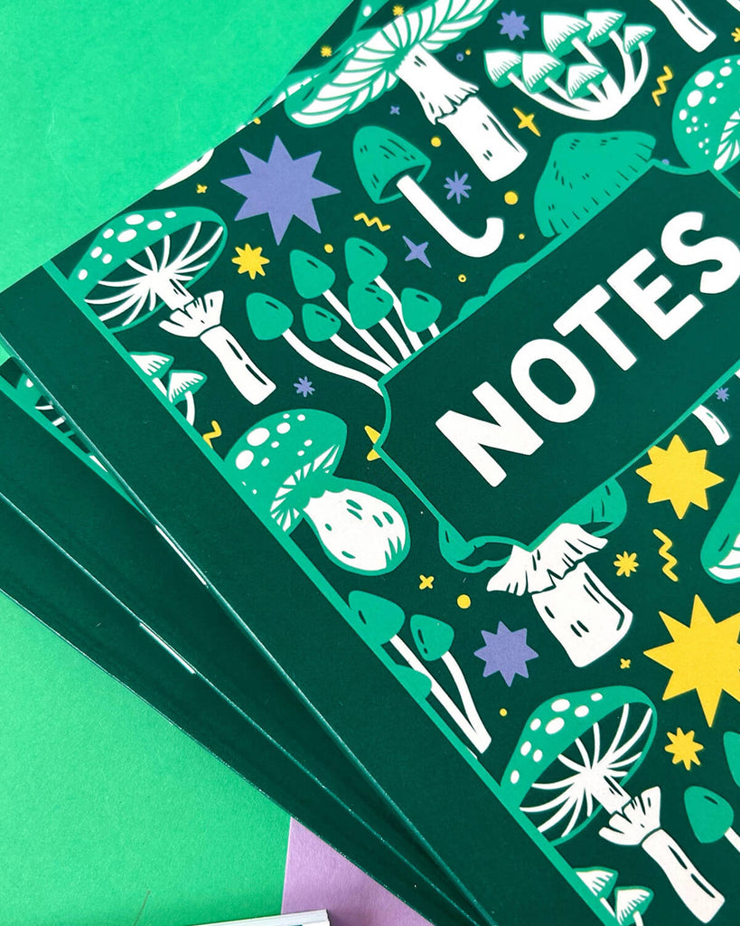 Close-up of the notebook cover showing detailed illustrations of mushrooms, stars, and toadstools in vibrant colours with the word “NOTES” in white on a dark green background.
