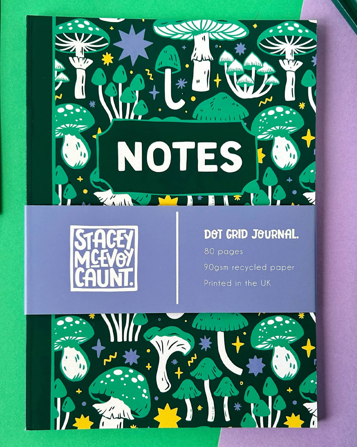 Full view of the Green Mushroom Dot Grid Journal, featuring a playful design of mushrooms, stars, and toadstools in green, lilac, and yellow tones, with a lilac band stating “Dot Grid Journal - 80 pages, 90gsm recycled paper, Printed in the UK.”