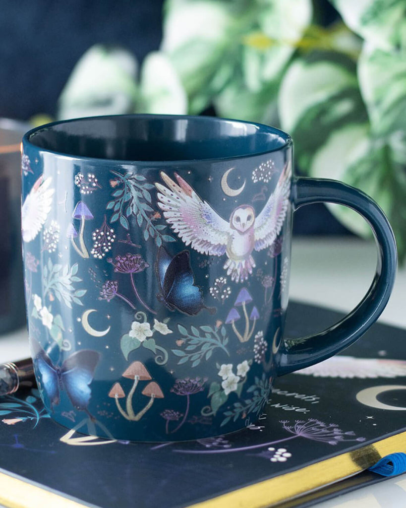 The mug styled on a table next to a lit candle and a decorative notebook. The mug’s rich, whimsical design contrasts with the soft candlelight, creating a cosy nighttime vibe.