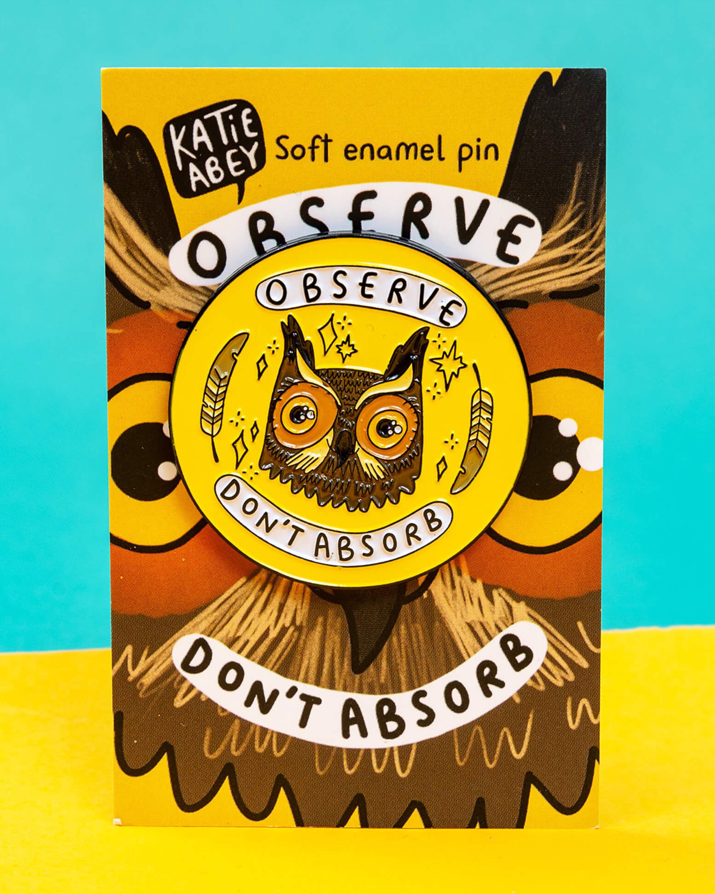 A Rebel Romance “Observe Don’t Absorb” enamel pin badge designed by Katie Abey, displayed on its illustrated backing card. The pin features a detailed owl face with feathers and stars on a bright yellow background, with the words “Observe” and “Don’t Absorb.” A quirky, empowering accessory celebrating alternative style for millennials.