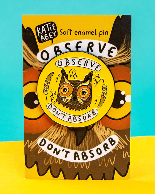 A Rebel Romance “Observe Don’t Absorb” enamel pin badge designed by Katie Abey, displayed on its illustrated backing card. The pin features a detailed owl face with feathers and stars on a bright yellow background, with the words “Observe” and “Don’t Absorb.” A quirky, empowering accessory celebrating alternative style for millennials.