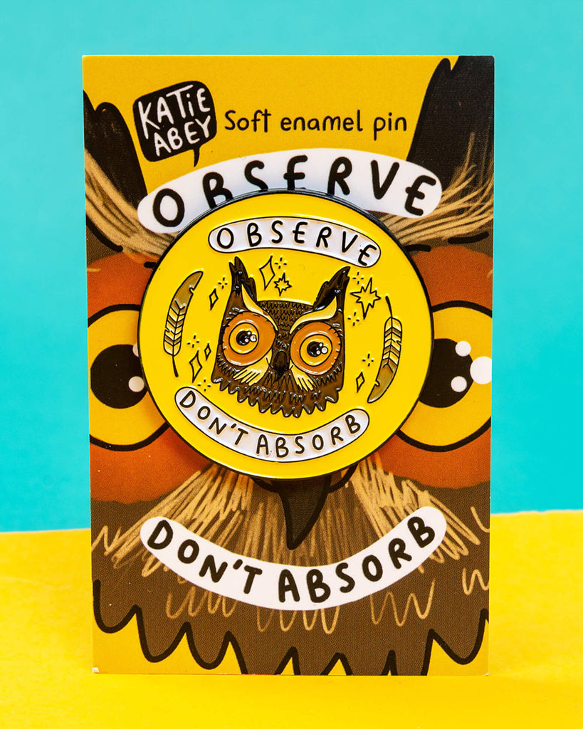A Rebel Romance “Observe Don’t Absorb” enamel pin badge designed by Katie Abey, displayed on its illustrated backing card. The pin features a detailed owl face with feathers and stars on a bright yellow background, with the words “Observe” and “Don’t Absorb.” A quirky, empowering accessory celebrating alternative style for millennials.