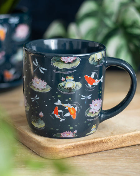 A close-up of a ceramic mug with a dark background and colourful koi fish, frogs, dragonflies, and water lilies, placed on a wooden coaster with plants in the background.