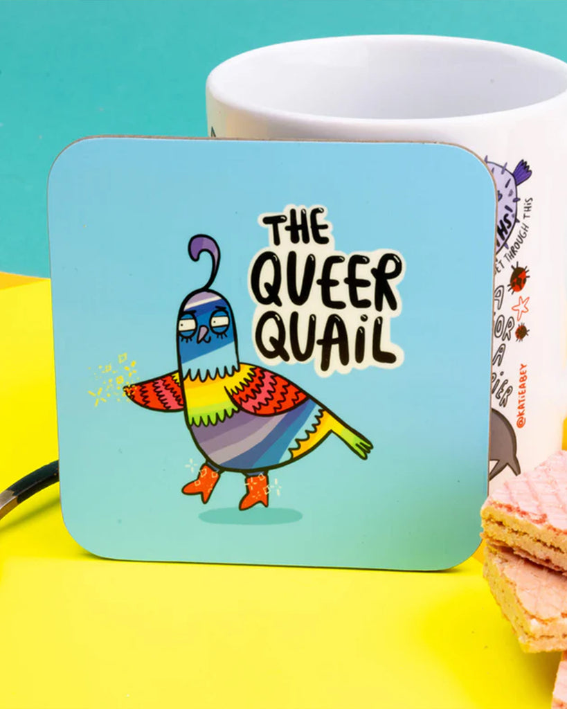 The Queer Quail coaster by Rebel Romance, designed by Katie Abey, shown on a bright yellow surface with a mug and wafers nearby. The colourful rainbow quail adds a playful, inclusive vibe to this alternative homeware from a UK brand known for celebrating unique aesthetics.