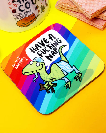 Katie Abey’s Nap Raptor coaster resting on a yellow surface next to a katie abey motivational mug and pink wafer biscuits. The rainbow design and text are clearly visible.