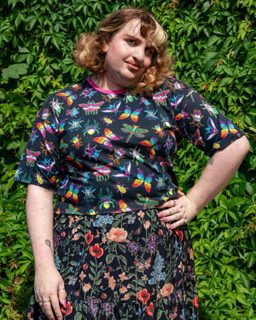 Maddie models the Rebel Romance OUTLET PRIDE Bugs Cropped Boxy Tee, featuring neon rainbow butterflies, beetles and dragonflies on black fabric. She stands with one hand on her hip against a leafy backdrop, showcasing inclusive alternative style from this ethical UK fashion brand for millennials.