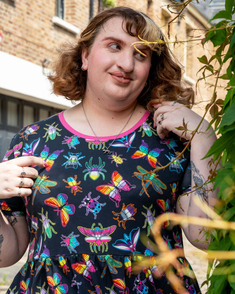 Maddie smiles playfully, touching her hair, wearing the Rebel Romance OUTLET PRIDE Bugs Stretch Smock Dress. The detailed rainbow insect print shines, perfect for millennial slow fashion lovers.